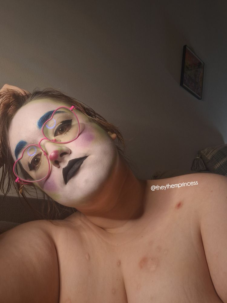 me looking down at the camera with my hand in my hair and wearing clown makeup (white foundation, green contour, purple cheeks, blue eyebrows, pink nose, black eyeliner and lipstick, lots and lots of highlight)