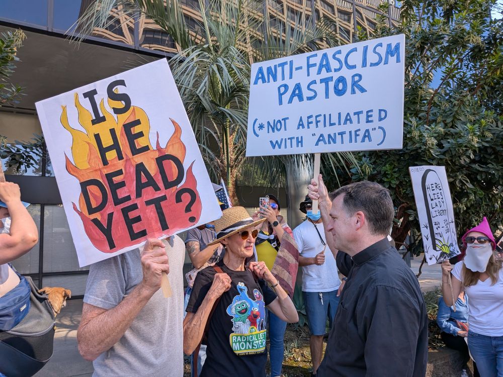 protesters holding signs including "is he dead yet?" over red, orange, and yellow flames and "anti-fascism pastor (not affiliated with "antifa"