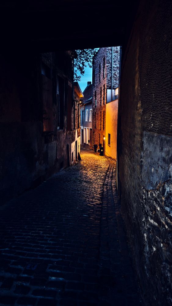 An arch leading into a tight alleyway, there's a bit of ambient light and one orange streetlight illuminating a portion of the street
