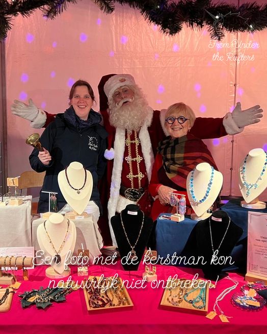 De maker van From Under the Rafters en haar schoonmoeder staan lachend naast de kerstman in de kraam, omringd door sieraden en winterse decoratie.