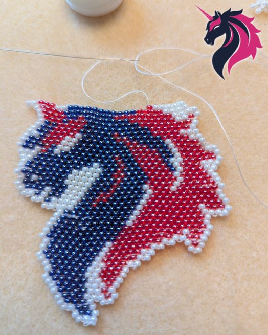 Close-up van het kralenlogo in de vorm van een eenhoorn. De kralen zijn roze, blauw en wit, strak naast elkaar geregen met fijn draad.