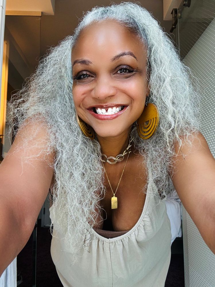 The author wearing a tan linen jumper, layered gold necklaces, & yellow wood earrings. Her white hair is pulled up in a half ponytail.
