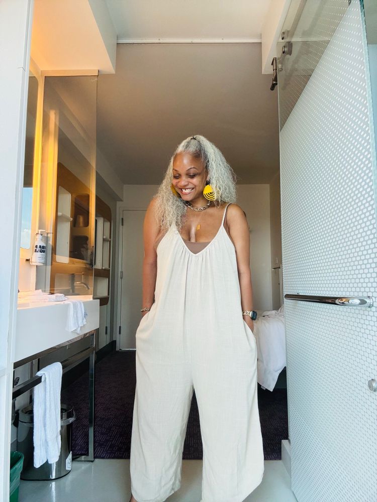The author wearing a tan linen jumper, layered gold necklaces, & yellow wood earrings. Her white hair is pulled up in a half ponytail.