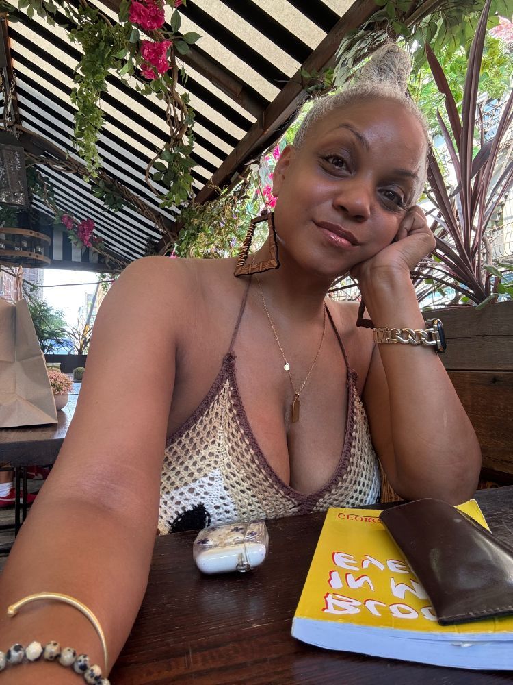 The author sits under a pagoda with pink flowers hanging from the rafters. She is having an yuzu matcha latte (out of frame,) & reading Blood in my Eye by George L. Jackson for Black August. She’s wearing a brown, tan, white and black crocheted top. A thin gold chain hangs from her neck with a Capricorn tarot card charm resting between her voluptuous breasts. Her wrist is adorned with a gold bracelet & a snowflake obsidian crystal bracelet for balance, purity, and grounding. Her other wrist sports an Apple Watch with a gold bracket strap. Her white and grey hair is pulled into a messy bun. Bamboo earrings made of wood with gold wire wrapping hang from her ears.