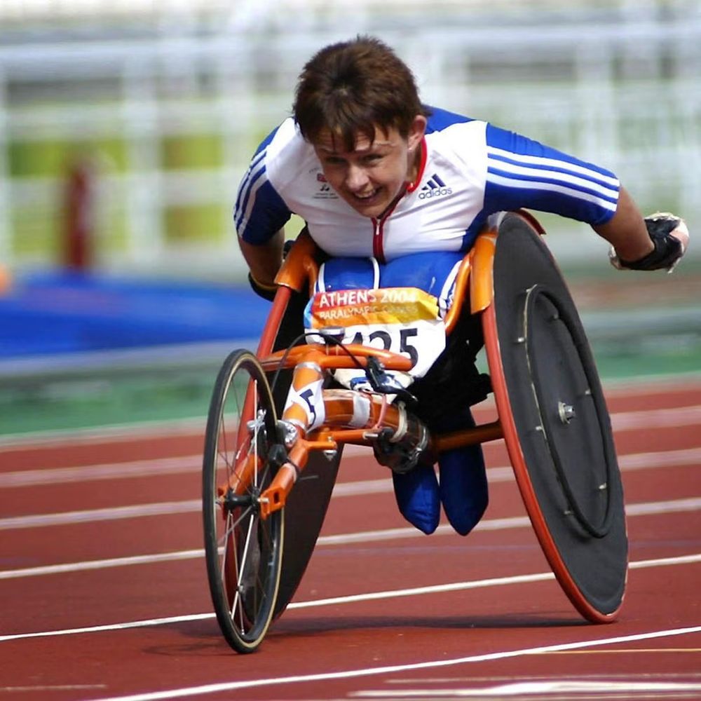 Tanni Grey-Thompson races in an orange wheelchair on a red track during the 2004 Athens Paralympic Games. Wearing a white and blue racing uniform, she leans forward mid-stride during a competitive event.