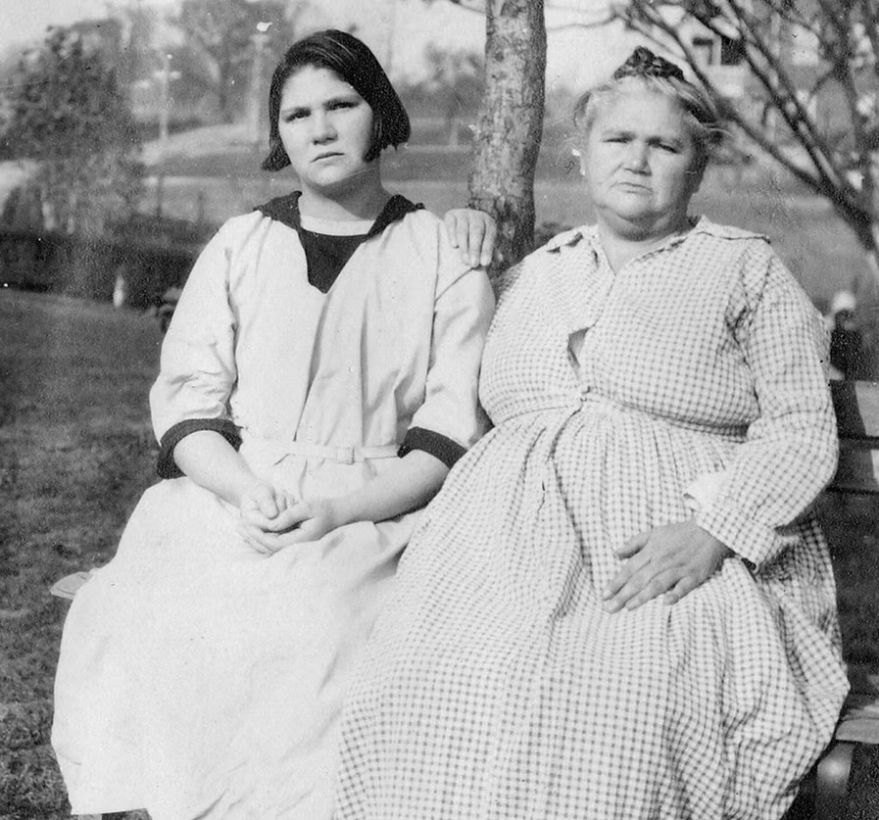 Black and white photo of a young woman, Carrie Buck, with short brown hair sits outside on a bench next to her mother, Emma Buck, whose light hair is pulled up. Both women wear long light-colored dresses and stare at the camera with furrowed eyebrows and serious expressions. Emma rests her right hand on Carrie's left shoulder.