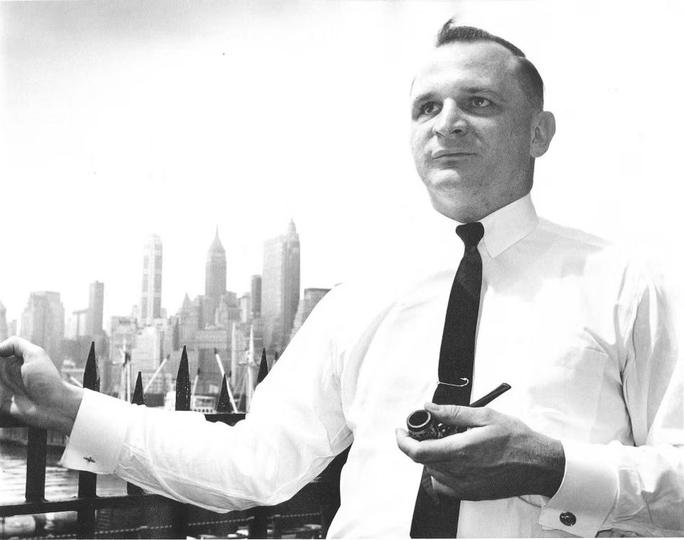 Robert Smithdas in a long-sleeved white collared shirt and dark tie holds a pie in his left hand and holds onto a spike metal fence. A city skyline is behind him.