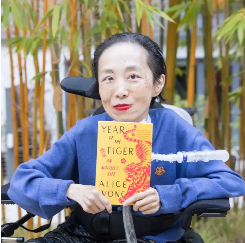 Alice Wong, an Asian American disabled woman in a power chair, against a background of bamboo trees. She is wearing a blue cardigan and sitting in a power chair. She is holding a copy of her memoir, Year of the Tiger, a paperback in yellow and red with a fierce tiger on it designed by Madeline Partner. She is wearing a bold red lip color and a trach at her neck. Alt text from Disability Visibility website.