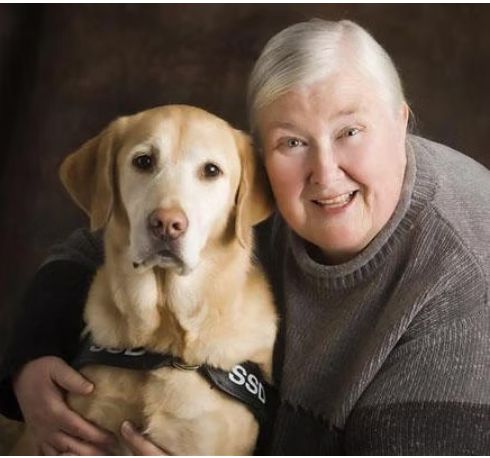 Sue Thomas with white hair wearing a dark sweater and smiling with her golden retriever.