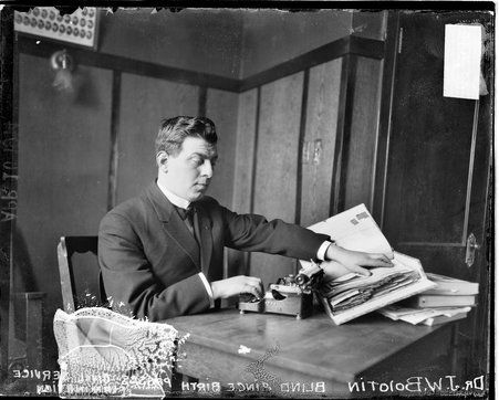 Half-length portrait of Dr. Bolotin sitting at a desk in a room, reading braille text with his left hand and typing with his right hand in a room.