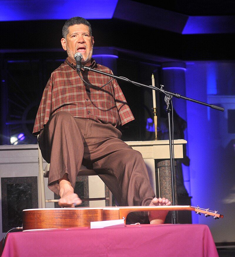 A seated Tony Meléndez sings at a microphone and plays a guitar with his feet.