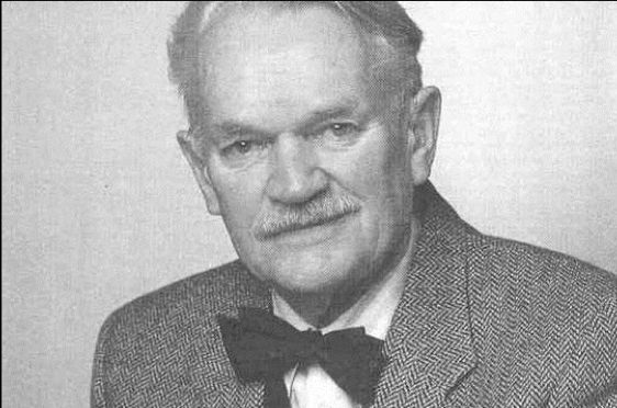 The image is a black-and-white portrait of an older Niels Erik Bank-Mikkelsen with a mustache, wearing a tweed jacket and a dark bow tie. He has a serious expression and is looking directly at the camera.