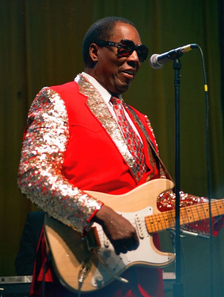 Clarence Carter wears sunglasses and a bright red vest over a gold-sequined jacket. He stands at a microphone holding a tan-colored guitar.