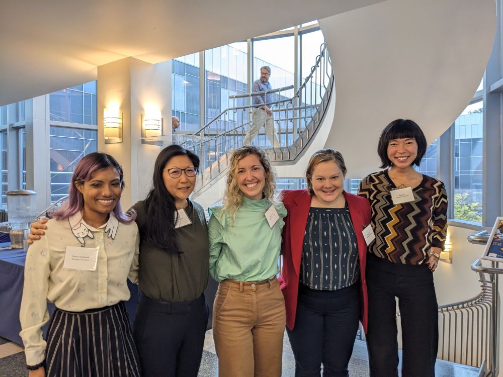 In a brightly lit law school hallway painted white, beneath a staircase spiraling upward, four Brooklyn Law students of Critical Race Theory and Work Law with professor Shirley Lin (second from left, woman, 40s, Chinese American, long black hair, metal dark rimmed glasses) shoulder to shoulder, all smiling.