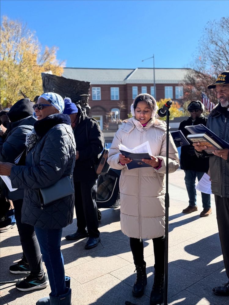 Photo of Aparna and a crowd of people, including veterans and descendants of Black veterans, reading into microphones outside
