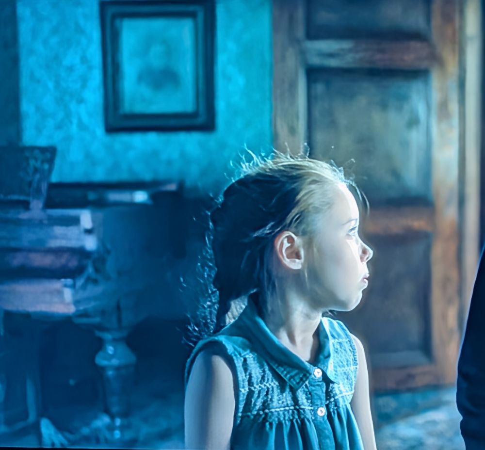Photo of episode 5 oh The Haunting of Hill House:
Young Nell in the foreground looking off the side to Theo. Behind her, the figure of a ghost can be seen faintly crouching under a piano, most visible by its white hands placed on the floor with fingers spread apart 