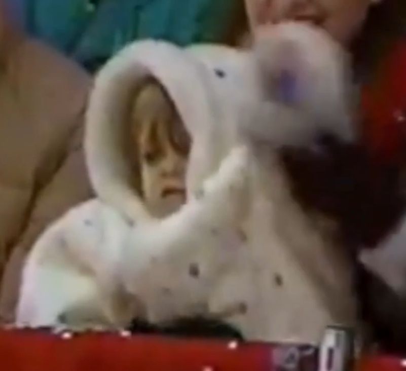 A very small child in a big fluffy winter coat looking confused as the adult they are with makes them wave at the Macy's Parade