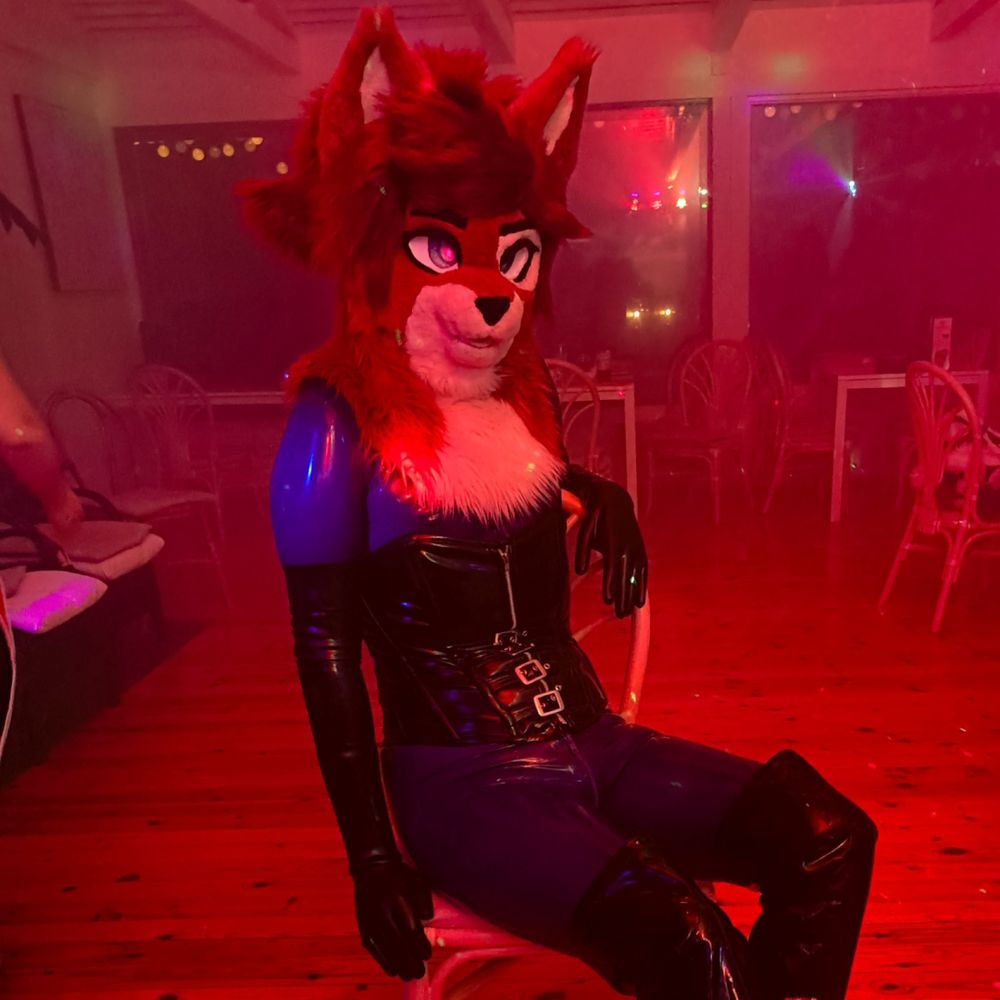 Fursuiter sitting on a chair giving teasing looks wearing blue shiny latex bodysuit, black latex gloves, PVC corset and boots.