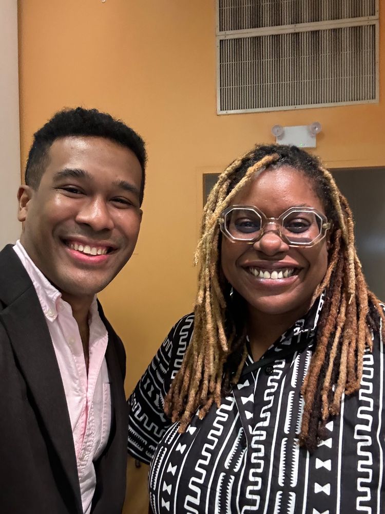 A Black Filipino man wearing a brown blazer and pink button up shirt takes a selfie with a Black woman wearing a black and white printed dress with honey brown locs