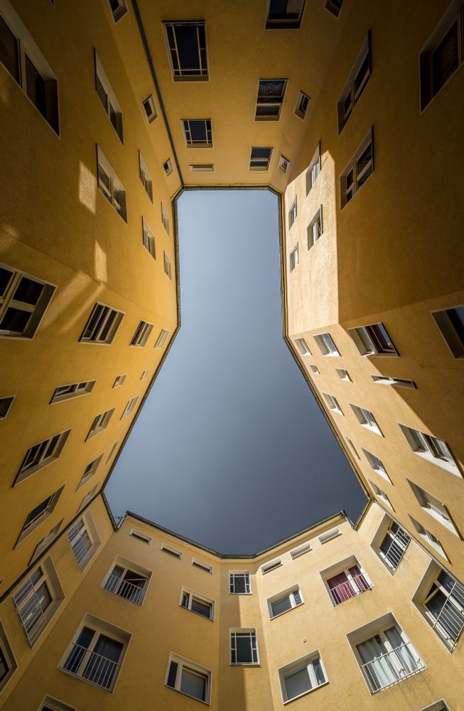 Das Bild zeigt den Blick nach oben aus einem Innenhof, der von einem mehrstöckigen Wohngebäude mit gelber Fassade umschlossen ist. Die Fenster sind regelmäßig und symmetrisch angeordnet, einige davon sind geöffnet oder mit Vorhängen und Rollos versehen. Der Innenhof öffnet sich nach oben in einem polygonalen Umriss. Der Himmel erscheint wolkenlos und in einem satten Blau. Sonnenlicht fällt von oben in den Hof und wirft klare Schatten der Fensterrahmen auf die gelben Wände.