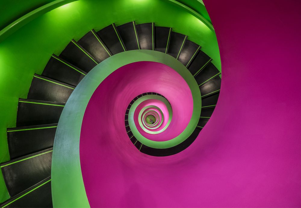 Der Blick von oben nach unten in eine kleine Wendeltreppe. Sie ist in hellgrün und pink gehalten. Die Stufen sind schwarz.