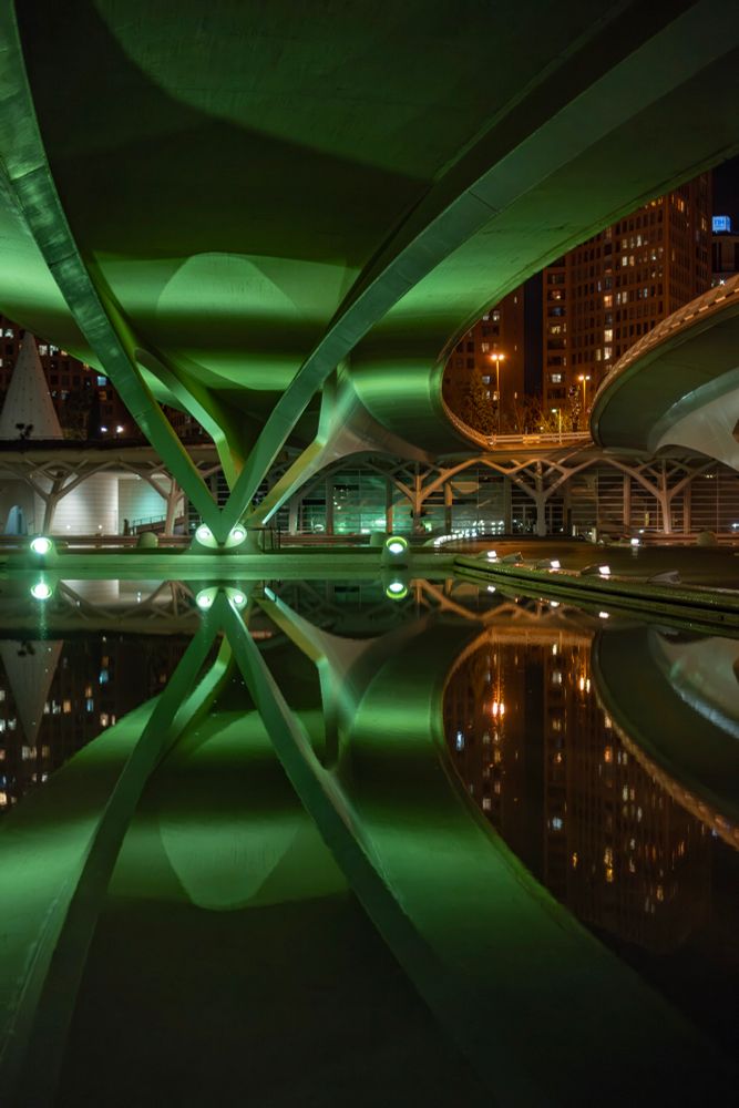 Das Bild zeigt eine nächtliche Aufnahme unter einer Brücke in der Wissenschaftsstadt von Valencia. Die moderne Betonstruktur ist durch grüne Lichtquellen eindrucksvoll beleuchtet. Die geschwungenen Träger und Säulen der Brücke formen ein geometrisches Muster, das sich symmetrisch in der ruhigen Wasserfläche darunter spiegelt. Im Hintergrund sind beleuchtete Gebäude der Stadt zu erkennen. Die Gesamtkomposition wirkt architektonisch komplex und fast futuristisch, mit starken Kontrasten zwischen Licht und Schatten.