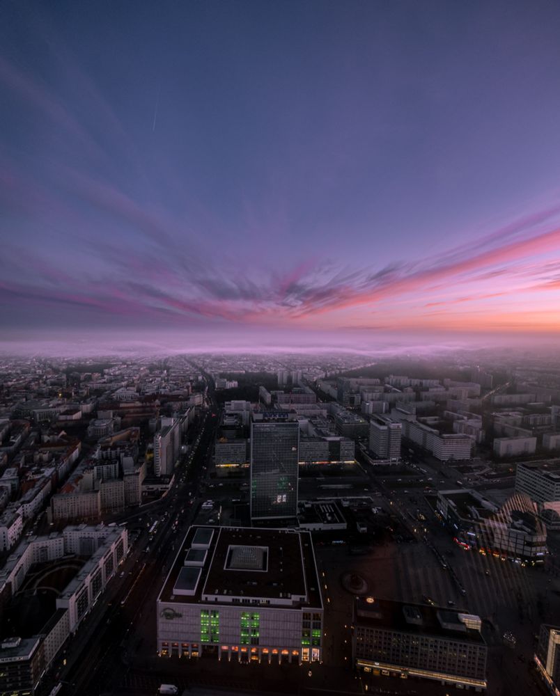 Das Bild zeigt einen atemberaubenden Blick aus dem Berliner Fernsehturm bei Sonnenaufgang. Die Stadt erstreckt sich unter einer sanften Nebelschicht, während sich der Himmel in violetten, rosa und orangefarbenen Tönen färbt. Langgezogene Wolkenstrukturen betonen die Weite des Himmels. Unten im Bild sind der Alexanderplatz mit dem markanten Hochhaus des Park Inn Hotels sowie das Einkaufszentrum Galeria zu sehen, dessen grün beleuchtete Fenster hervorstechen. Die Straßen der Stadt beginnen sich mit Verkehr zu füllen, während die Morgendämmerung Berlin in ein sanftes Licht taucht.