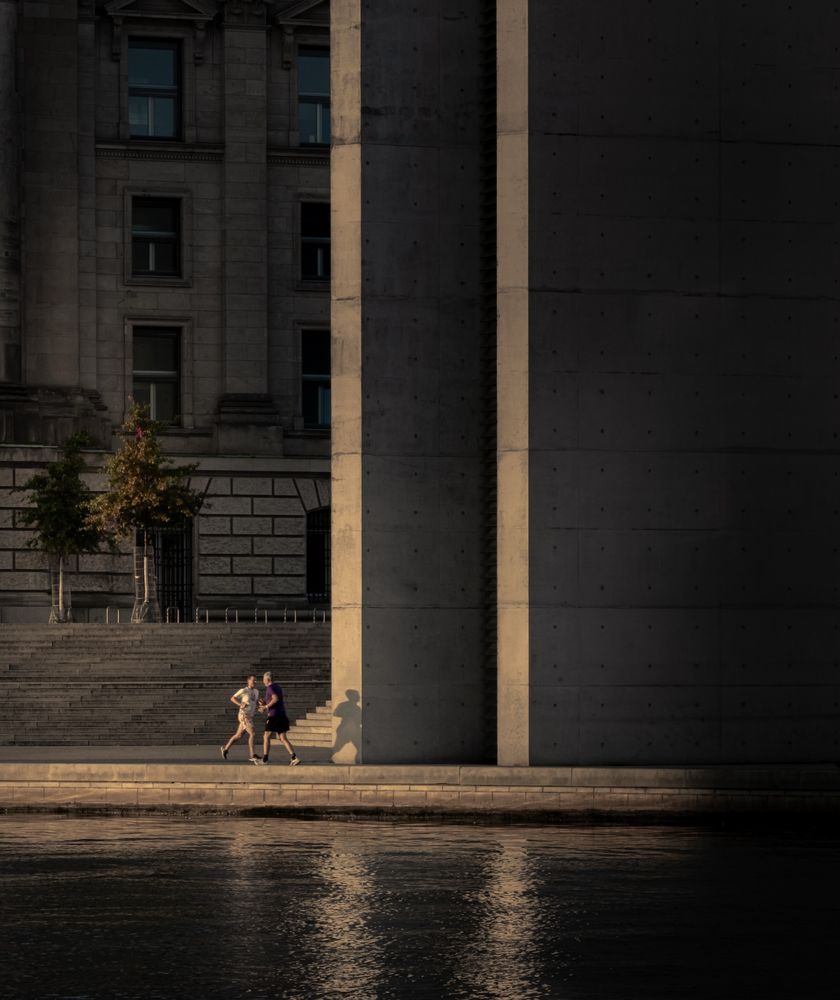 Das Bild zeigt zwei joggende Personen am Ufer eines Flusses in einer städtischen Umgebung. Sie befinden sich auf einem schmalen Weg direkt am Wasser, der von großen, modernen Betonsäulen flankiert wird. Dahinter ist ein historisches Gebäude mit einer Fassade aus Naturstein und mehreren rechteckigen Fenstern zu sehen. Zwei kleine Bäume stehen vor dem Gebäude, und Treppen führen zu einer erhöhten Ebene. Die Szene ist in warmes, seitlich einfallendes Licht getaucht, das lange Schatten auf die Wand und den Boden wirft. Das Wasser im Vordergrund spiegelt das Licht leicht.
