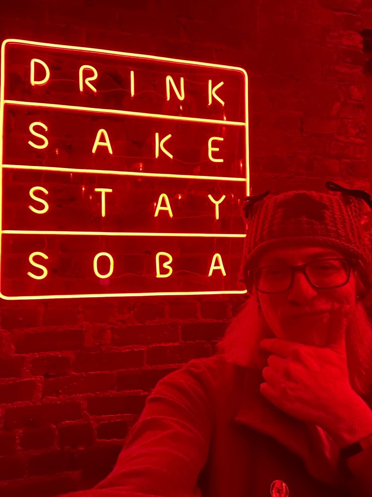 A photo of a woman bathed in red light, making a silly face in front of a neon style sign that reads, “Drink Sake Stay Soba.”