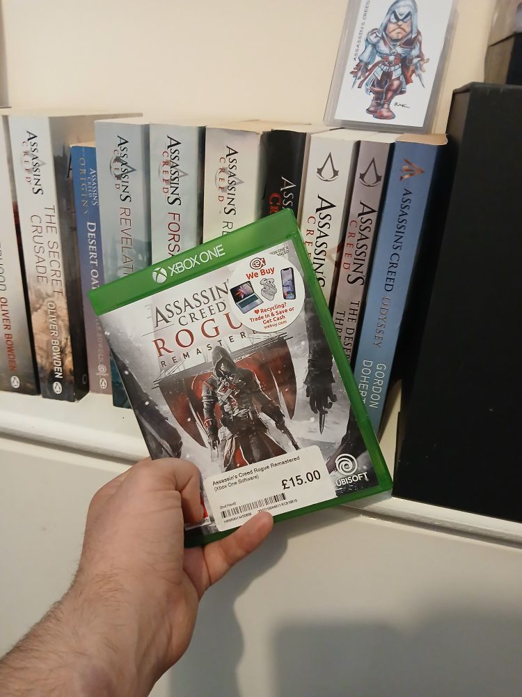 A picture of me holding the xbox One case for Assassins Creed rogue remastered. The case has a green edge with the Assassins Creed Rogue title on the center. The case also has a picture of shay cormac with his ship in the background.

There is also 2 stickers on the gane case on in the top right that has the CeX logo one in the bottom center that shows its price as £15 