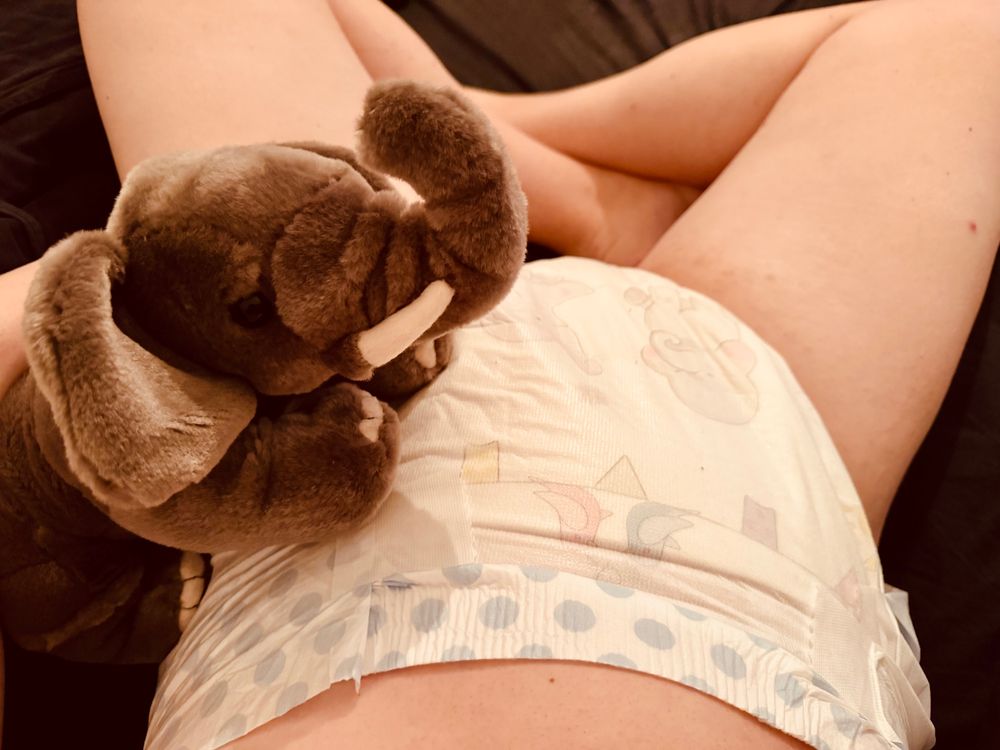 A person lying on a bed wearing a diaper with an elephant pattern, holding a plush elephant toy.