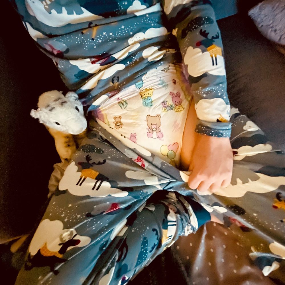 A person wearing festive blue pajamas with reindeer and cloud patterns is sitting on a couch. They are partially pulling down the waistband of their pajamas to reveal a diaper with colorful baby-themed illustrations, including teddy bears and balloons. A small white plush toy rests beside them.