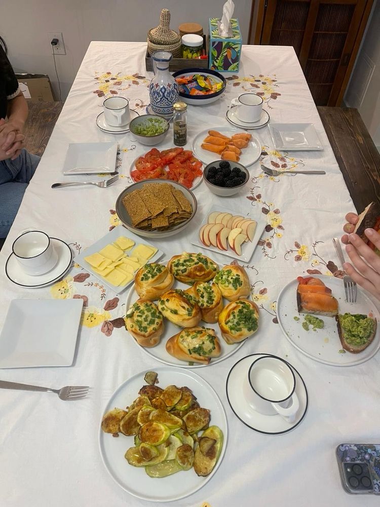 A white table cloth with zucchini, scallion bread, apple, sourdough crackers, tomato, and other food items on white plates. There are also smaller eating plates scattered around the table.