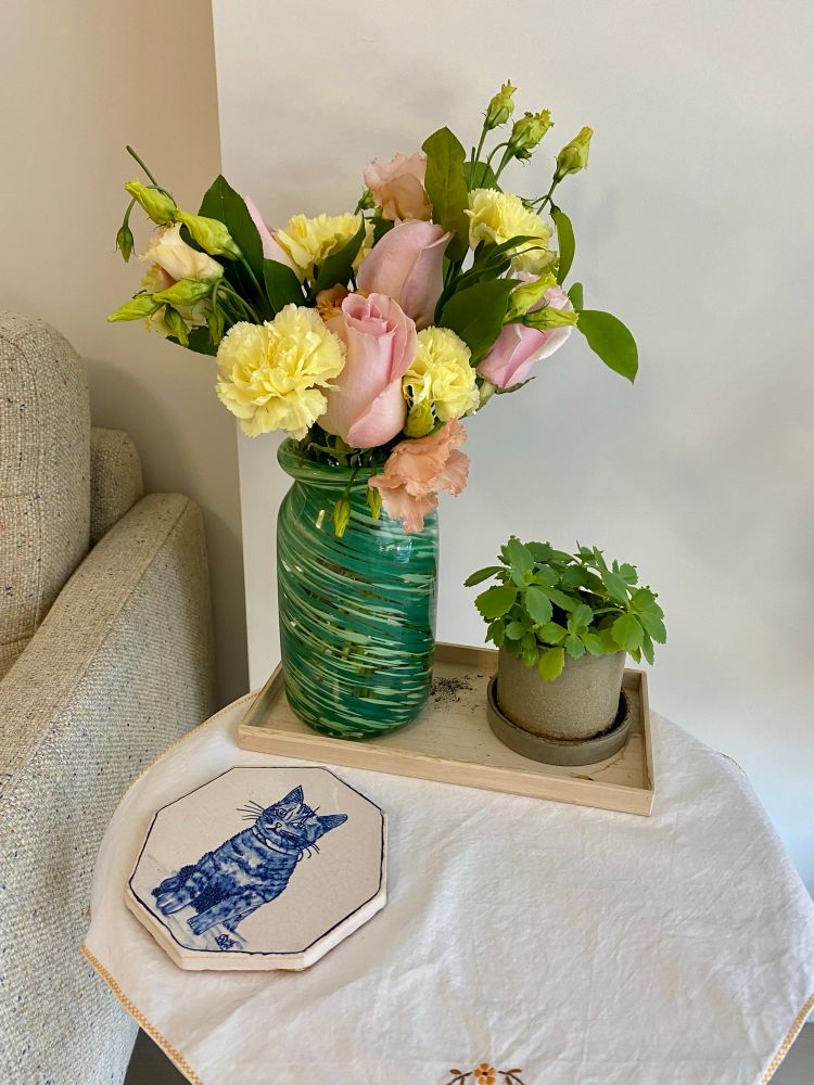 A little side table with a green vase filled with yellow and pink flowers. On the right of the vase is a smaller plant. 