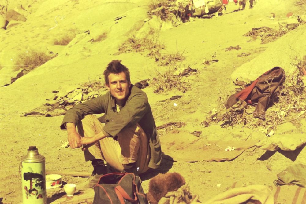 Young English man wearing nondescript clothing sitting down before a cup, bowl and thermos flask, smiling at the camera. Photograph circa 1 March 1985.