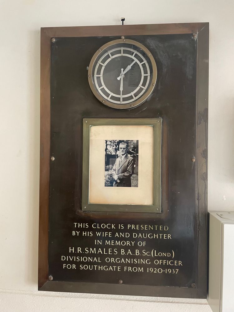 A picture of a very old clock framed in some sort of brass plate, with a black and white photo of a man underneath. The text reads: “this clock is presented by his wife and daughter in memory of H.R. Smales B.A B.Sc (Lond) Divisional Organising Officer For Southgate from 1920-1937”