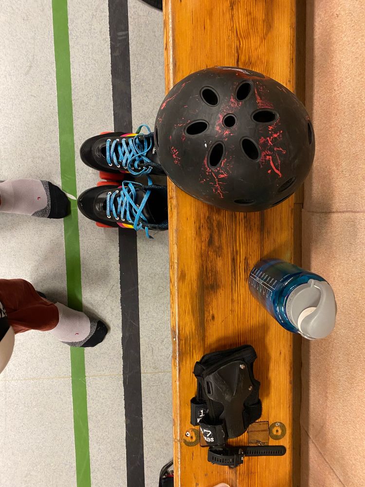 Mes pieds en chaussettes, mes rollers (quads noirs à roues rouges, lacets bleus à paillettes, et détails arc en ciel sur les côtés), et sur un banc mon casque noir et traces de peinture rouge, ma gourde, et mes protections poignets