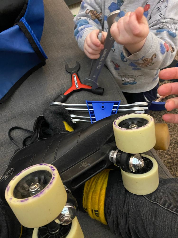 POV assise sur le canapé, un patin quad de roller derby sur les jambes. Mon bébé me tend ma clé de 16. Il y a d’autres outils sur le canapé dont un en forme de Y spécifiquement pour les patins 