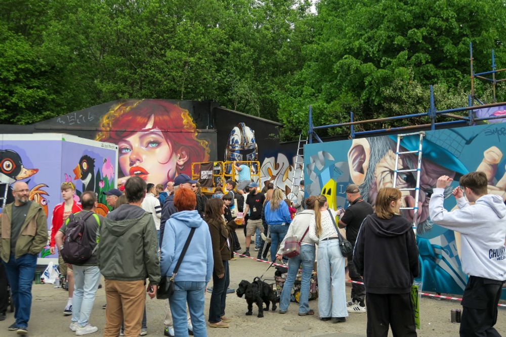 Photo of the crowds at Yardworks, Glasgow in SWG3. In the background are several large work in progress murals depicting colourful ducks, a red haired woman, an angry man in a blue hat.