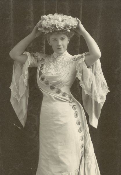 A photograph of the poet Ella Wheeler Wilcox, a white woman wearing a white dress and holding a wreath garland on top of her head.