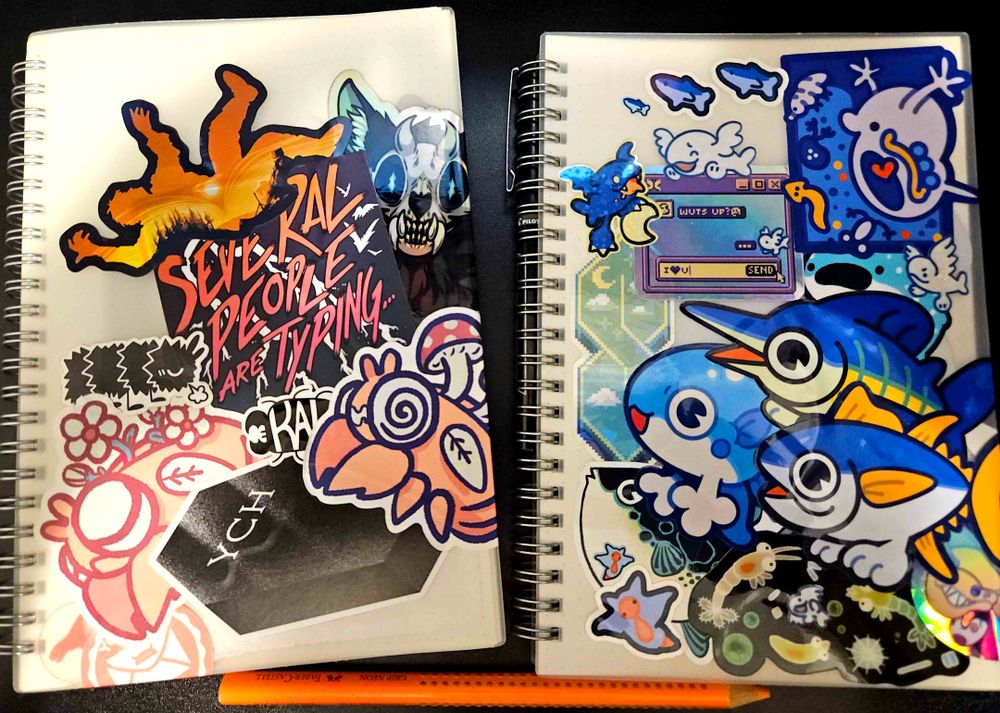 Two sketchbook or notebooks sit side by side. Both are covered in various stickers. The one on the left leans towards an orange color theme, while the one on the right is blue with fish. 