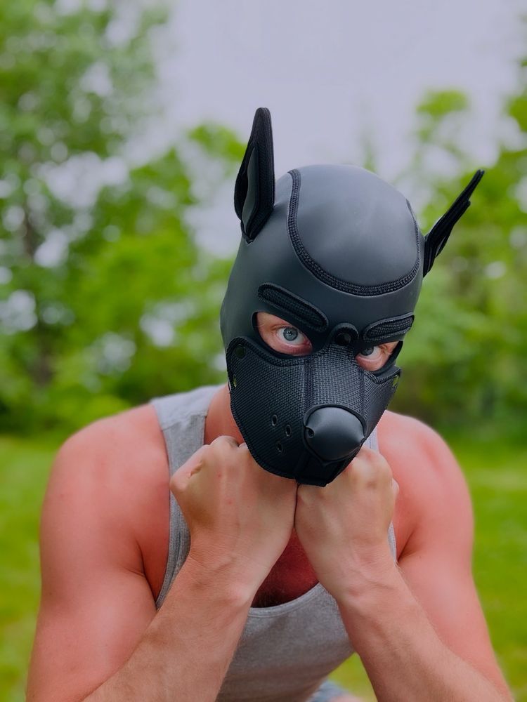 A portrait of me outside wearing a black pup hood and a grey tank top. My chin is resting on my knuckles and there is green grass and trees in the background 