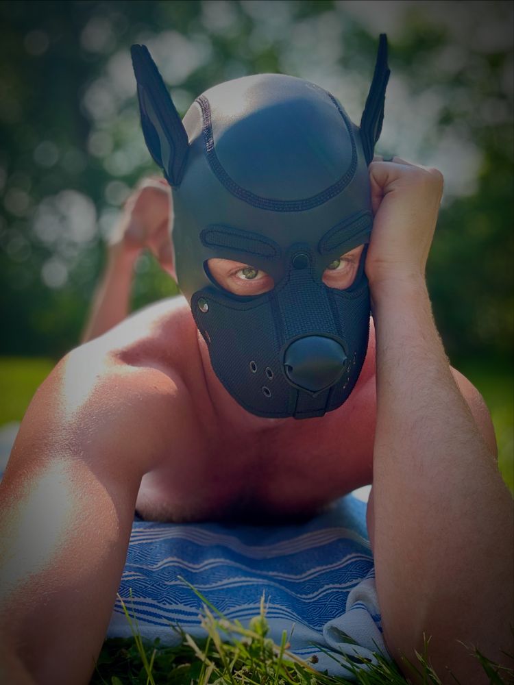 Me laying on my stomach in the grass on a sunny day on a blue towel wearing a black pup hood taking a selfie. I’m shirtless and barefoot with feet in the air and head resting on my left hand