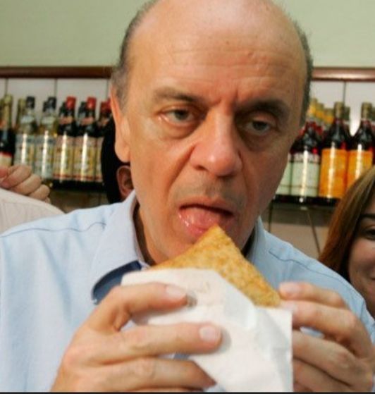 Foto de José Serra, um homem branco e careca, comendo um pastel com cara de nojo.