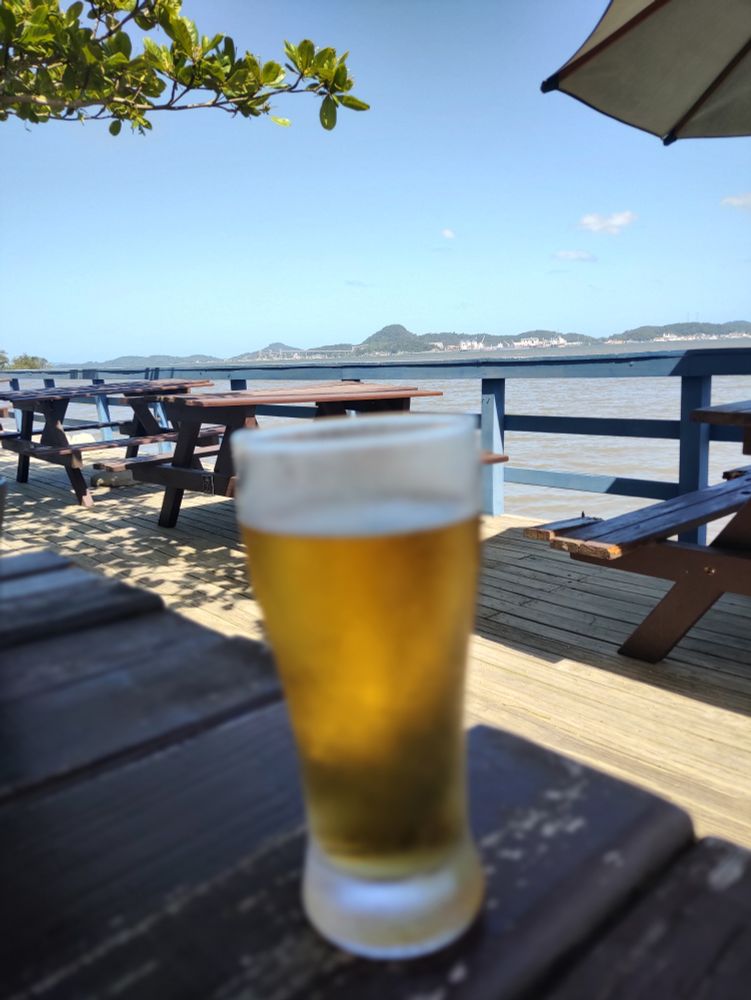 Mesa de madeira sobre um deck. Ao fundo o mar da Baia de Babitonga. O céu está azul, sem nuvens, as construções que aparecem são da cidade de São Francisco do Sul.