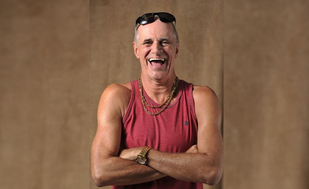 Foto do personagem Leleco, da novela Avenida Brasil. Um homem branco, magro, careca, com um largo sorriso. Ele veste camiseta regata vermelha, usa um relógio dourado, um colar de ouro e óculos escuros na testa.