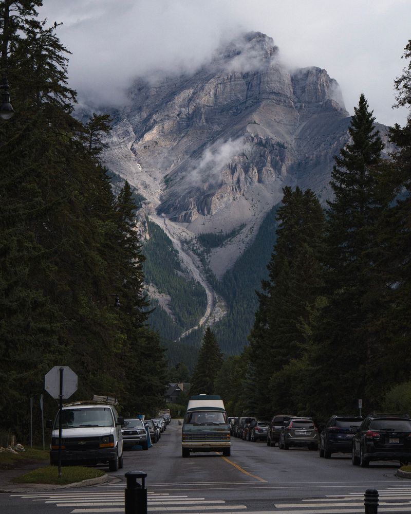 Looking down a street at a cloud covered mountain in the background, cars parked along the street, and a vintage van in the foreground.