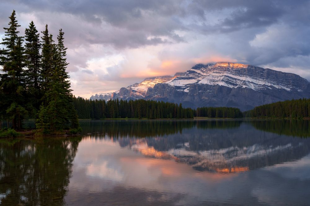 Mountain lit by sunrise golden light being reflected by a forested lake.