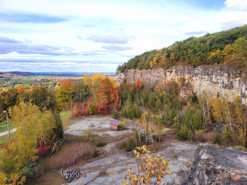 Kelso Conservation Area - a cliff with green and yellow trees is on the right, above a flat rocky area surrounded by trees with red and yellow fall colours. There is a small building in the center with a maroon colored roof. The sky is cloudy with patches of blue 