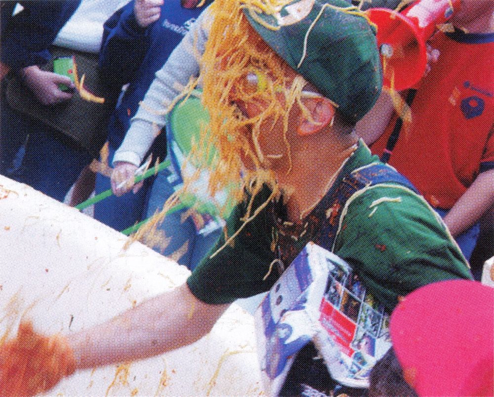 An action shot of a man dressed like Luigi getting drenched with a faceful of spaghetti. He's holding some kind of Gamecube swag bag.